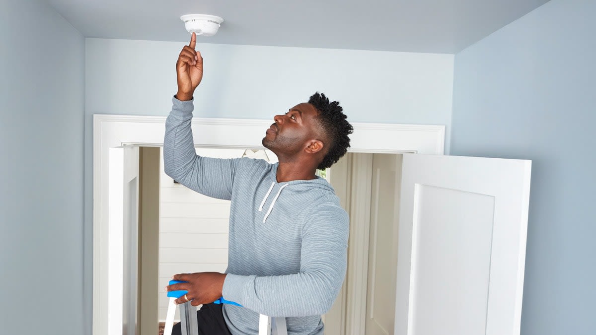 Man testing a ceiling-mounted carbon monoxide alarm using a ladder.
