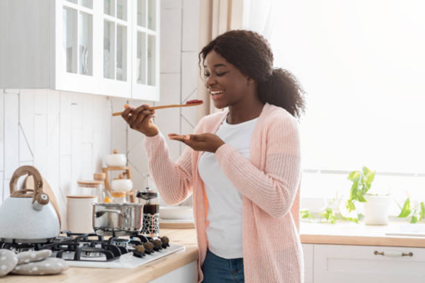 A woman stands in a bright kitchen tasting food from a wooden spoon while cooking on a stovetop with pots and a kettle nearby.